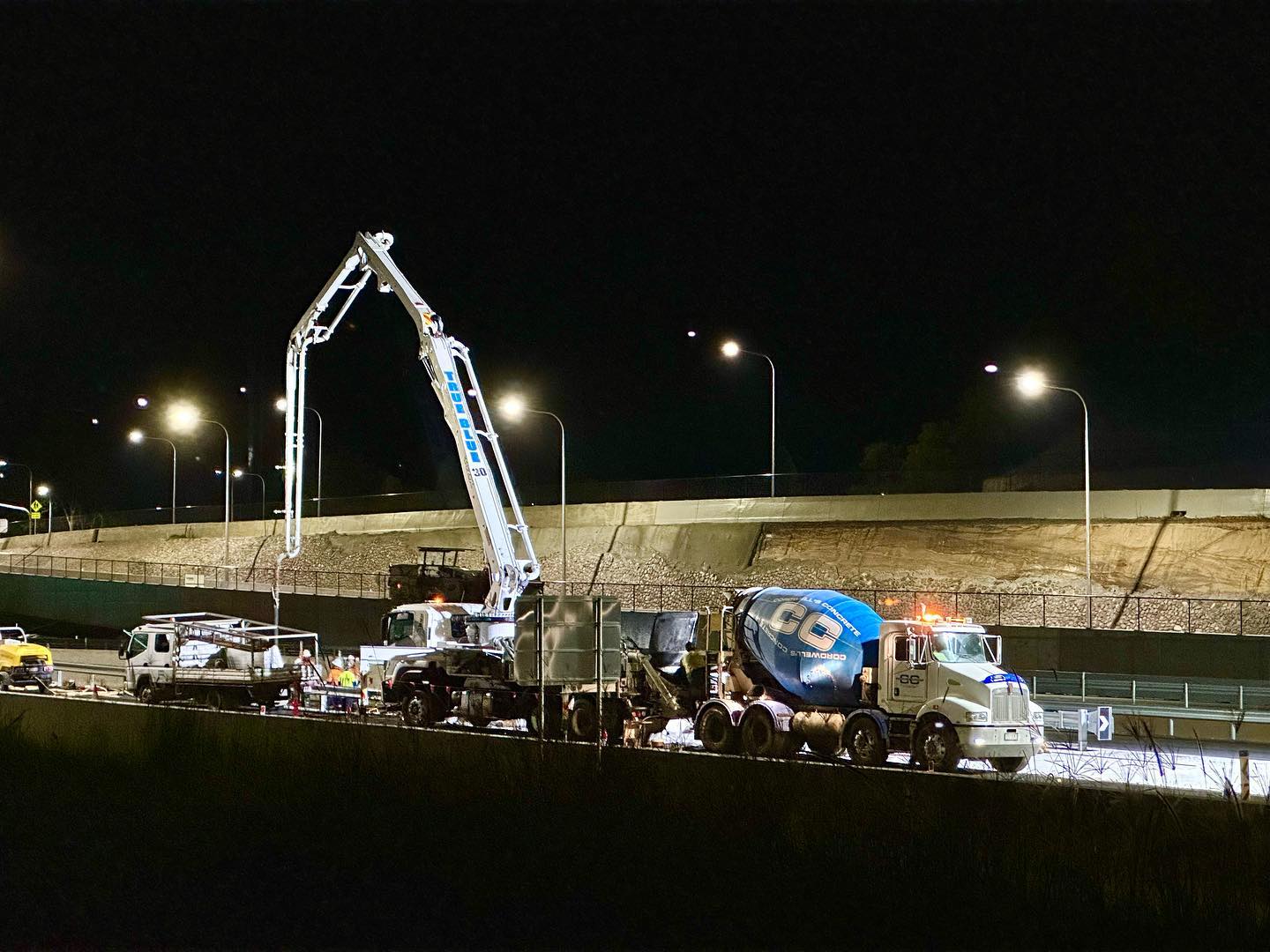 A construction truck is actively working on a highway at night, surrounded by darkness and illuminated by its headlights.  